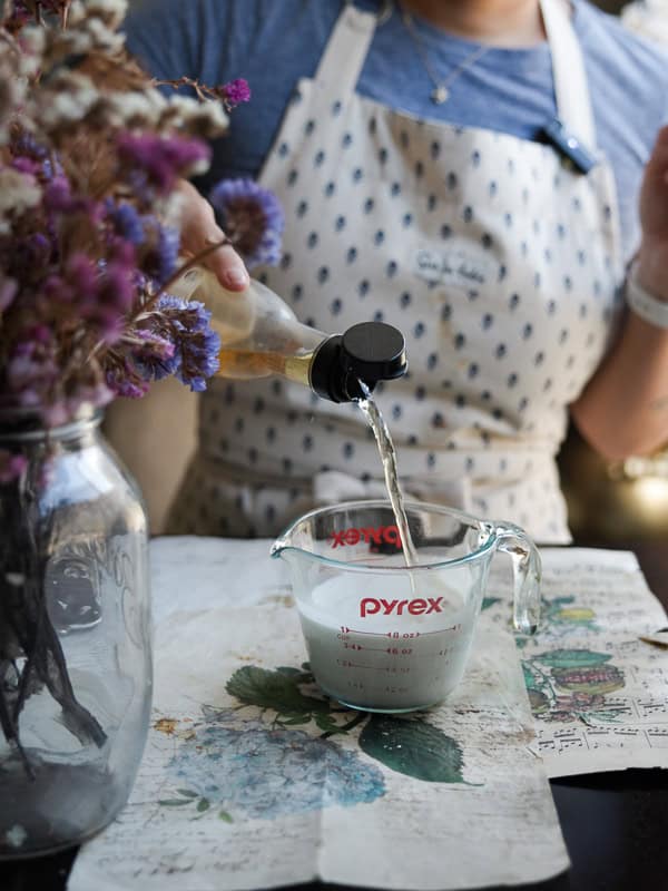 A person wearing a patterned apron pours liquid from a bottle into a Pyrex measuring cup filled with oat milk. Flowers in a glass jar are visible on the left. The scene is set on a table with floral paper.